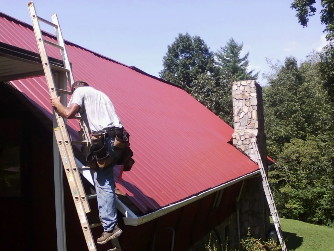 Roofer on a ladder beside a red metal roof and stone chimney — Soffit & Fascia Repair by Roofing Brothers in Burgin